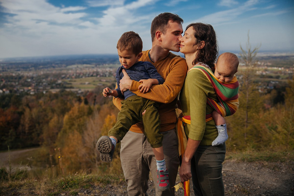 fotografia rodzinna- rodzice stoją blisko siebie, mężczyzna całuje żonę, dzieci są blisko rodziców